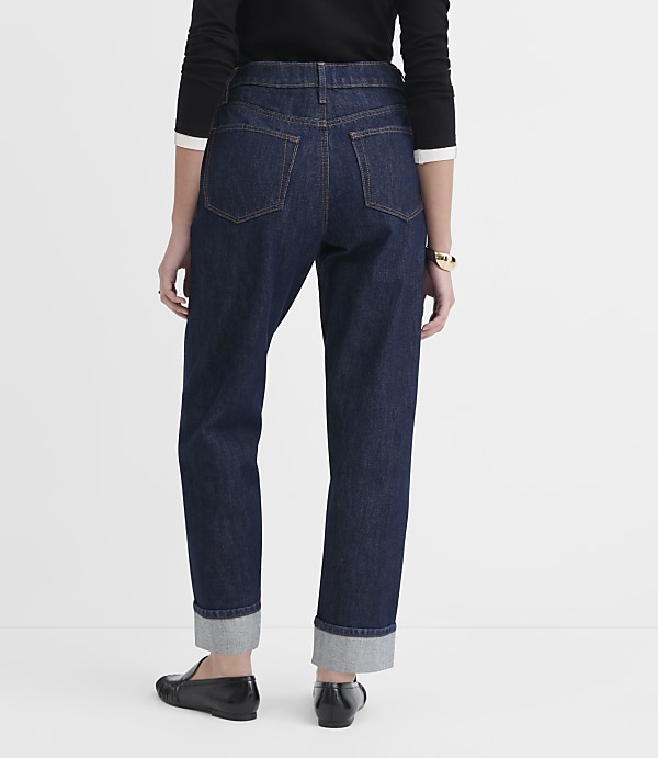 A person wearing dark wash cuffed jeans and black loafers stands against a plain white background.