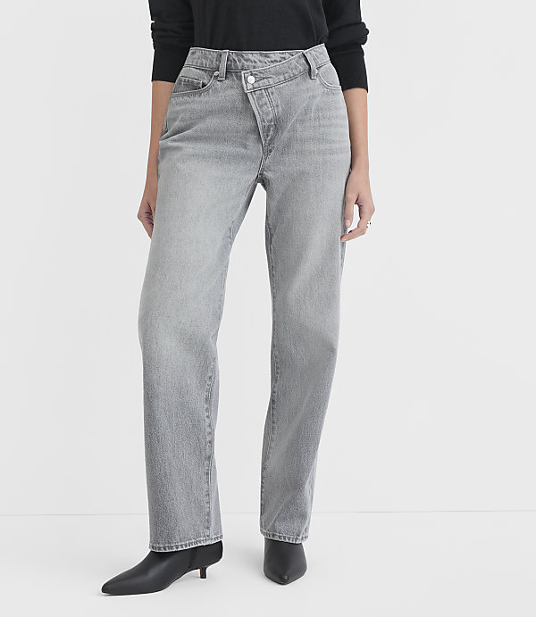 A person models light gray, asymmetrical-waist jeans and black pointed-toe boots against a plain white background.