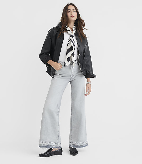 A woman in a black leather jacket, white t-shirt, and light-wash wide-leg jeans stands against a plain white background.