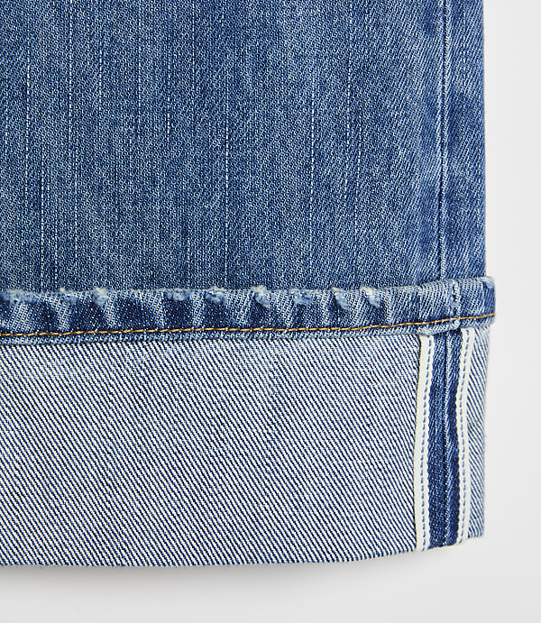 A close-up of cuffed blue denim jeans with a white selvedge edge is presented against a plain white background.