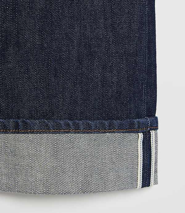 A close-up of dark blue denim jeans with a cuffed hem revealing a lighter blue selvedge edge against a plain white background.