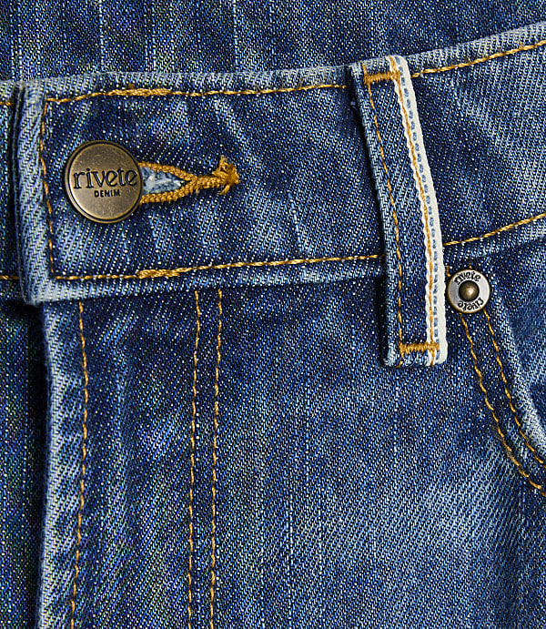A close-up of blue denim jeans features a brass button with "rivete DENIM" on the left and a small metal rivet on the right, with yellow stitching throughout.