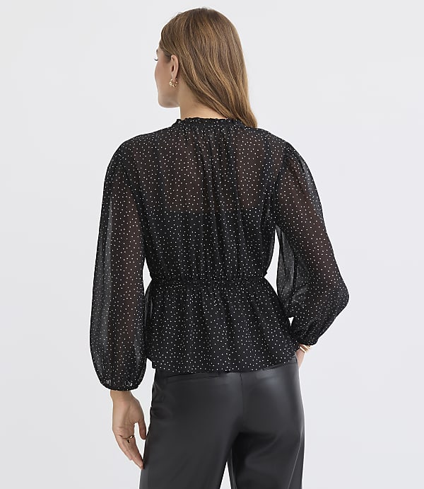 A woman in a black polka dot blouse and leather pants stands against a plain white background.