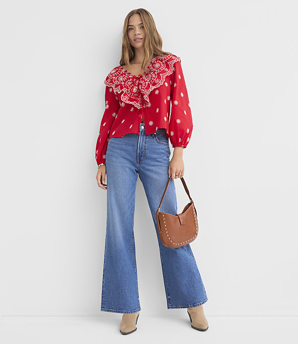 A woman in a red ruffled blouse and wide-leg jeans stands against a plain white background, holding a brown studded purse.