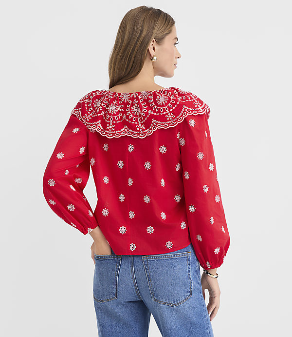 A woman in a red embroidered blouse and blue jeans stands against a plain white background.