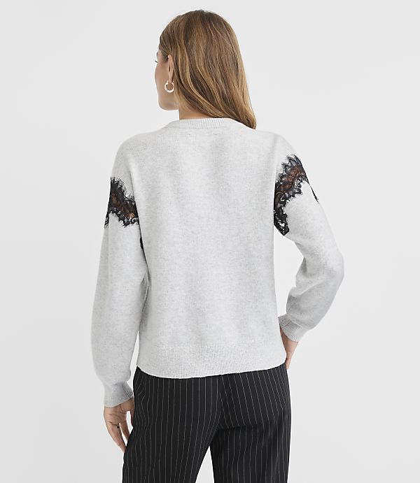 A woman in a light gray sweater with black lace accents on the shoulders and pinstripe pants stands against a plain gray background.