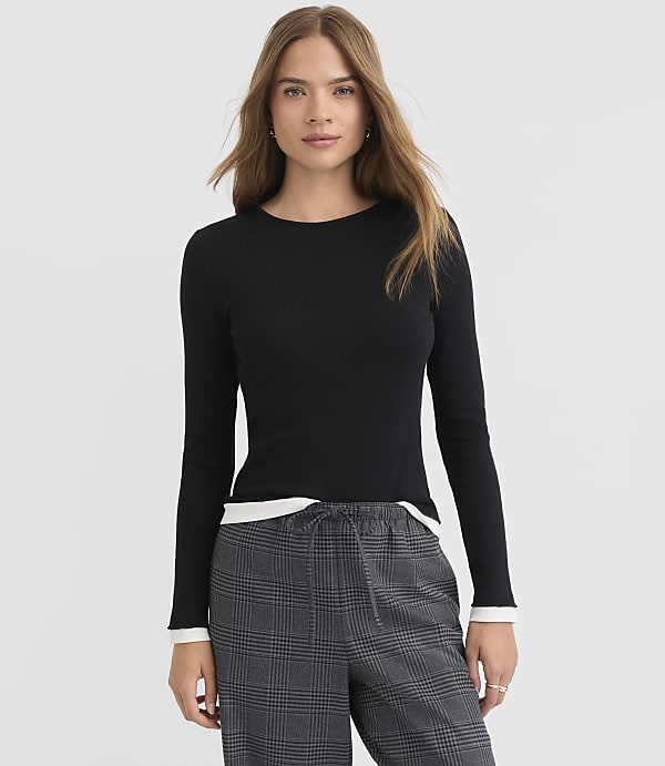 A woman in a black long-sleeved shirt and plaid pants stands against a plain white background.