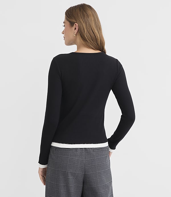 A woman in a black sweater and plaid pants stands against a plain gray background.