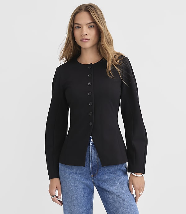 A woman in a black jacket and blue jeans stands against a plain white background.