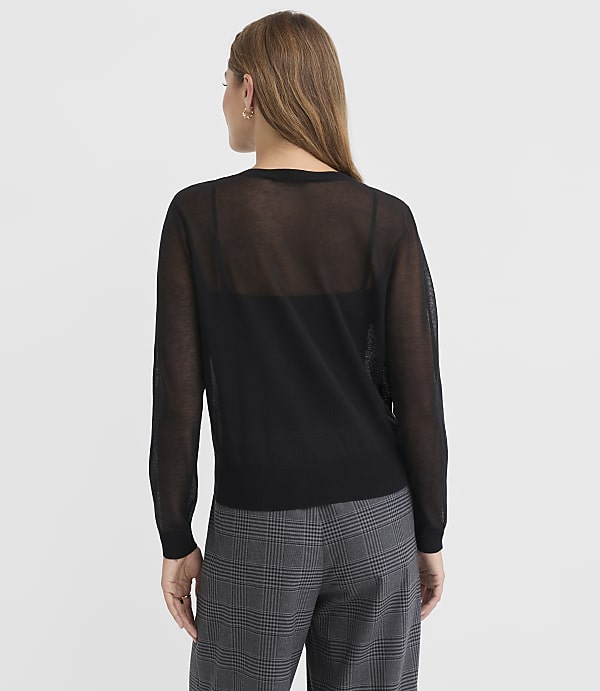 A woman in a sheer black sweater and plaid pants stands against a plain white background.