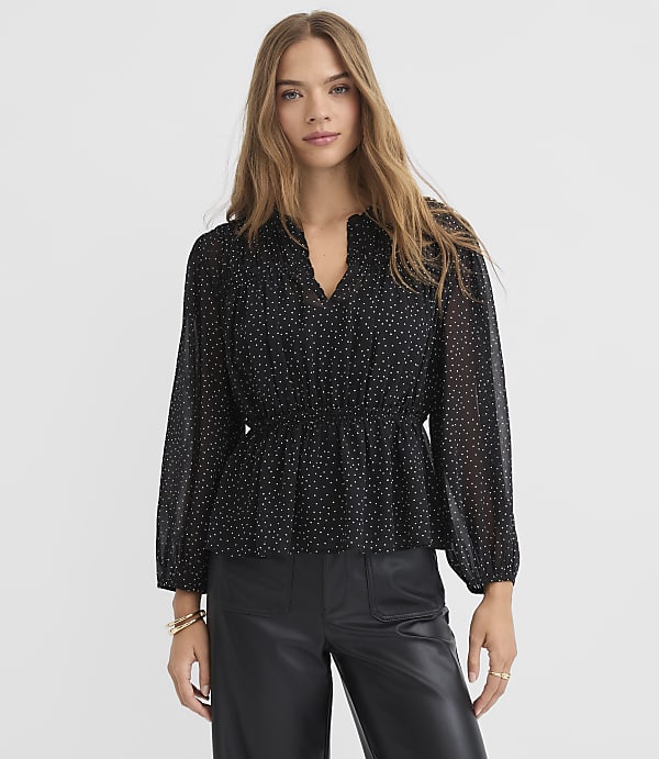 A woman in a black polka dot blouse and leather pants stands against a white textured background.