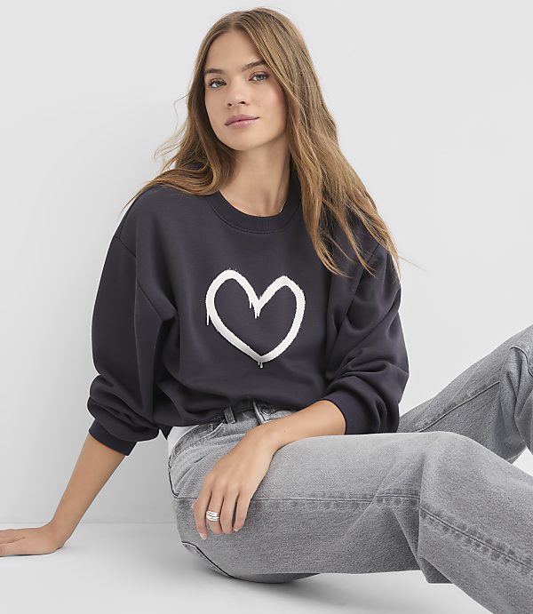 A young woman in a dark sweatshirt with a white heart graphic and grey jeans sits against a plain white background.