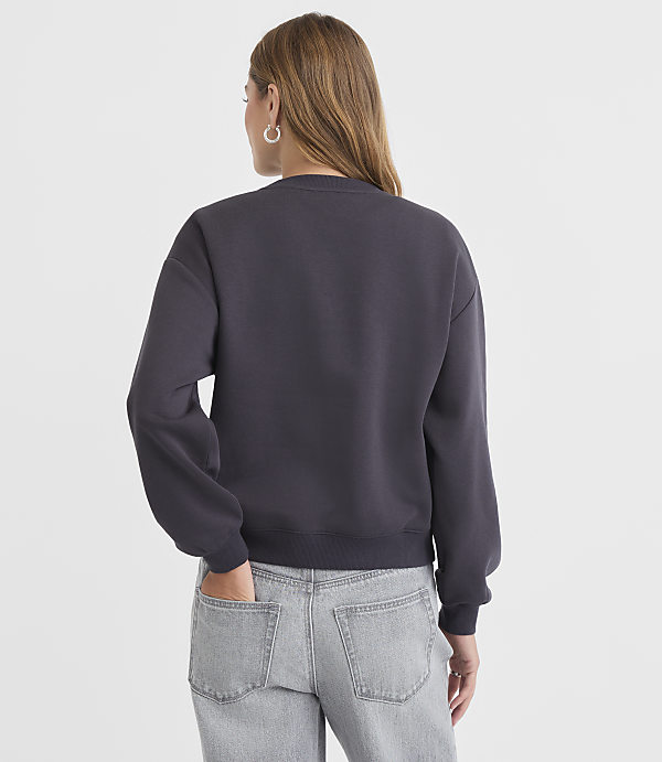 A woman in a dark gray sweatshirt and light gray jeans stands with her back to the camera, her left hand in her pocket, against a plain white background.
