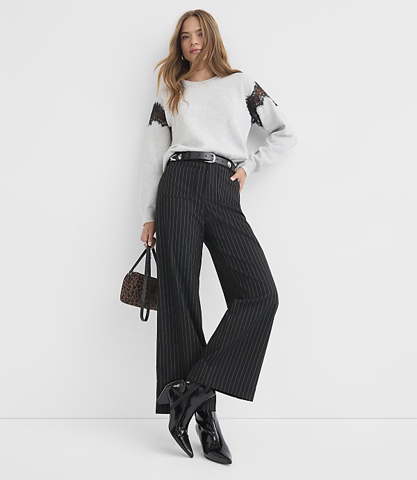A woman in a light gray sweater and pinstripe pants stands against a plain white background, holding a small patterned bag.