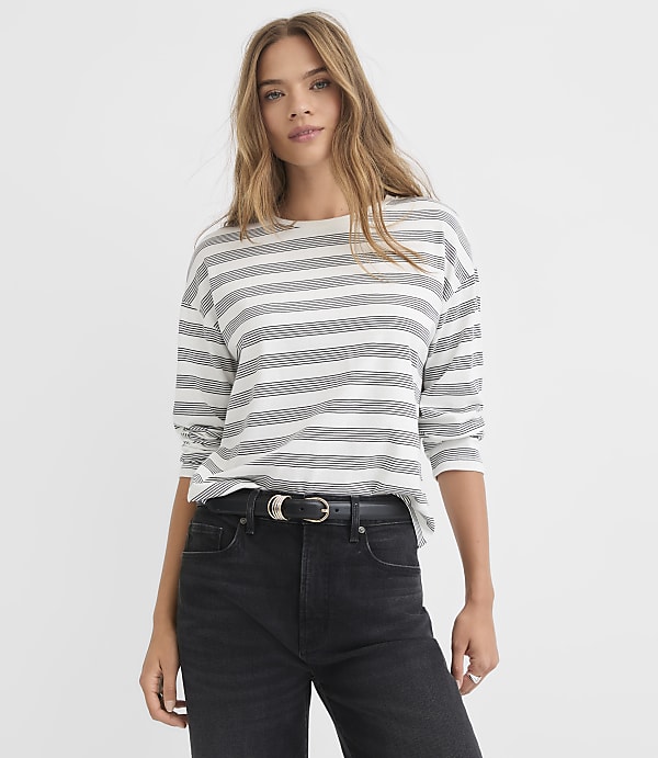 A woman in a striped shirt and black jeans stands against a plain white background.