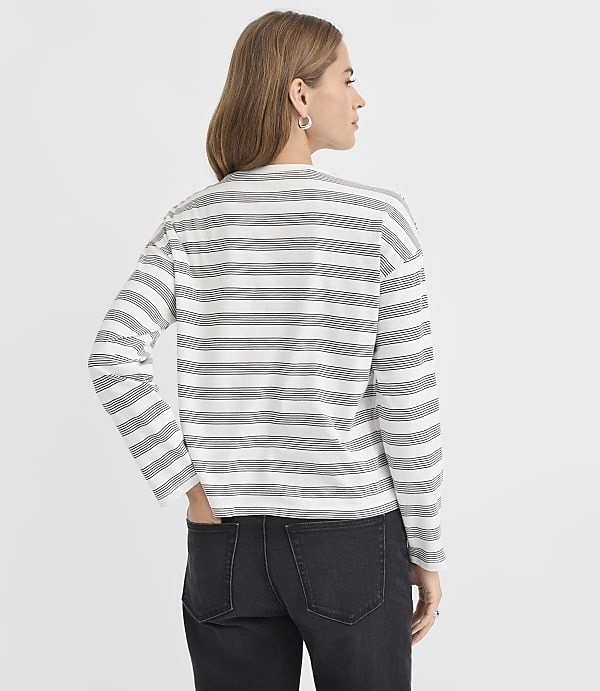 A woman in a striped long-sleeved shirt and dark jeans stands against a plain white background.