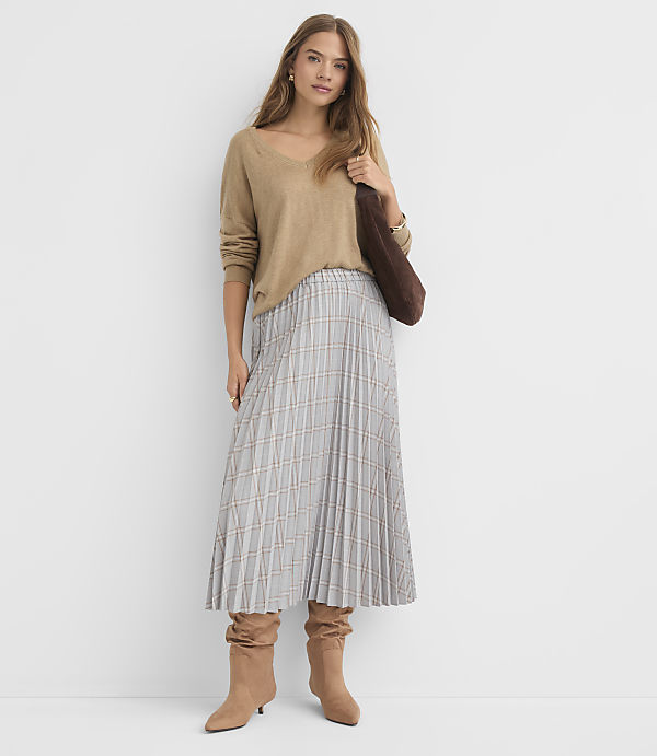 A woman in a tan sweater and plaid skirt stands against a plain white background, holding a brown bag.