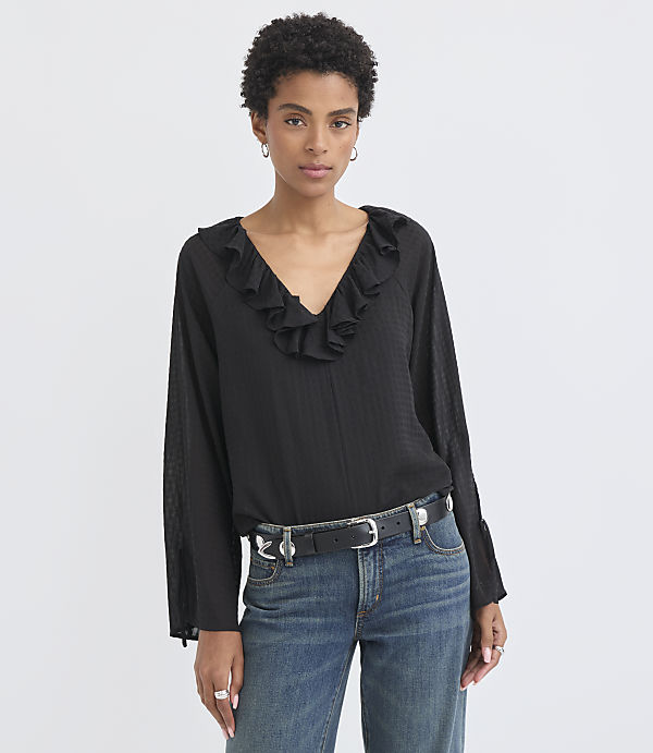 A woman in a black ruffled blouse and jeans stands against a plain white background.