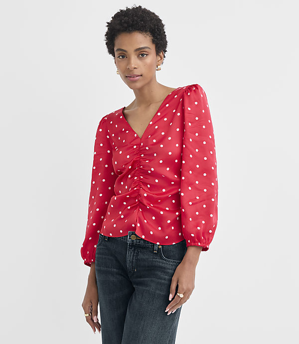 A woman in a red polka dot blouse and dark jeans stands against a plain white background.