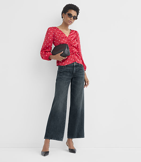 A woman in a red polka-dot blouse and wide-leg jeans stands against a plain white background, holding a black clutch.