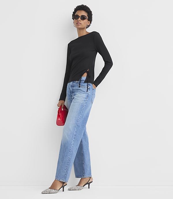 A woman in a black top and blue jeans stands against a plain white background, holding a red clutch.