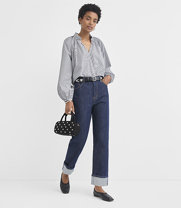 A woman in a striped blouse and dark jeans stands against a plain white background, holding a small black purse.