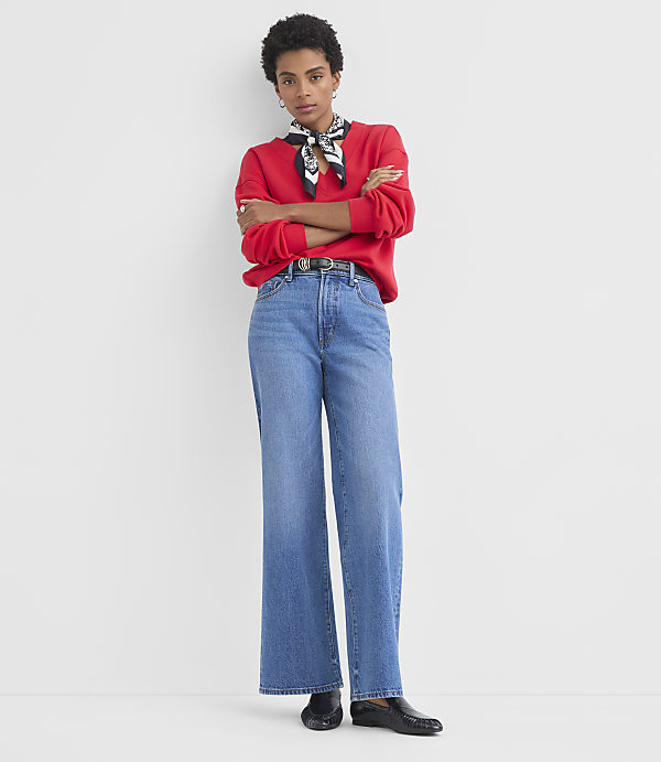 A woman in a red sweatshirt and wide-leg jeans stands with her arms crossed against a plain white background.