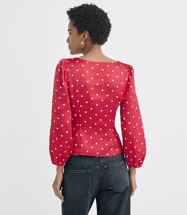 A woman in a red polka dot blouse and dark jeans stands against a plain white background.