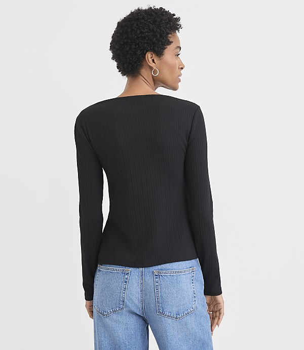 A woman in a black ribbed long-sleeved shirt and blue jeans stands against a plain white background.