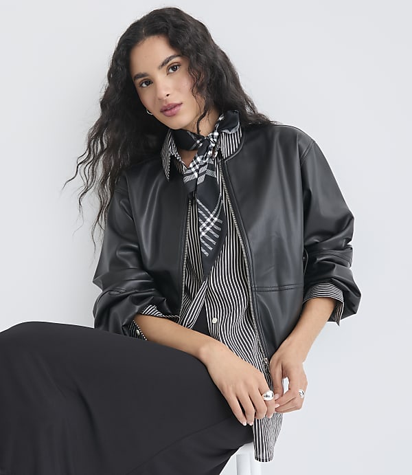 A woman with dark, wavy hair and a patterned scarf over a striped shirt and leather jacket sits against a plain white background.