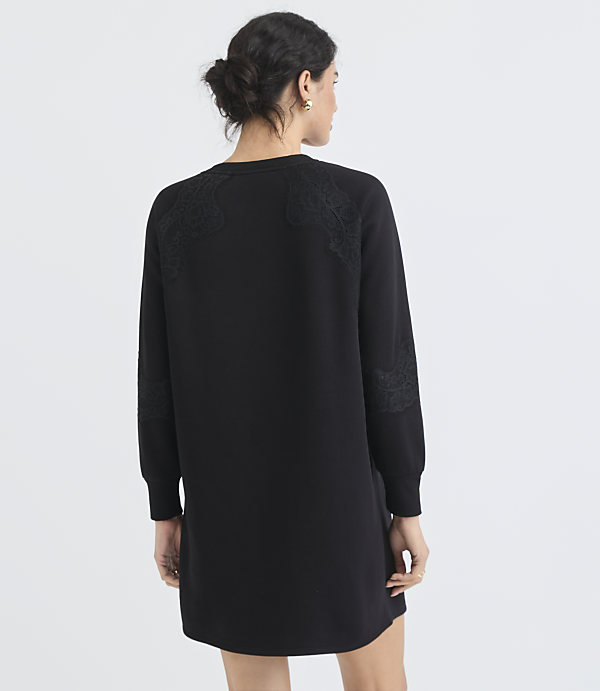 A woman in a black dress with lace detailing on the shoulders and sleeves stands against a plain white background.