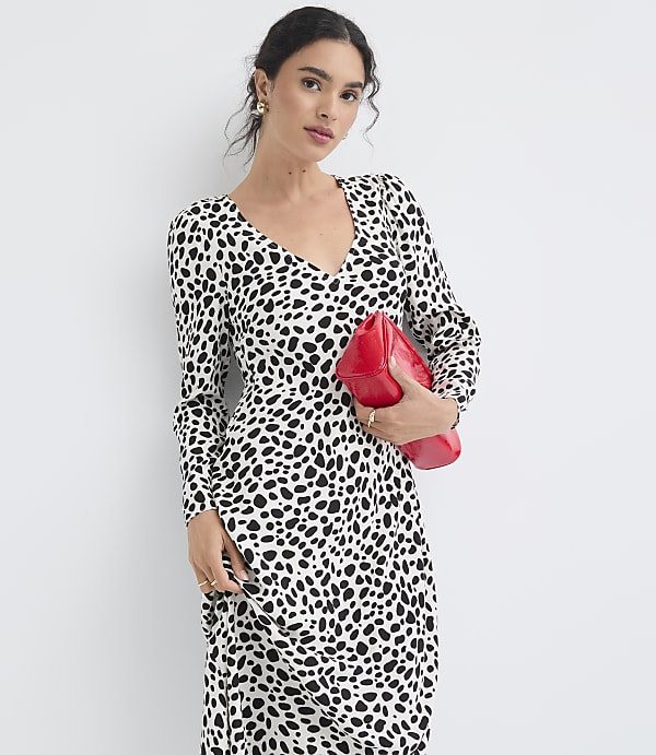 A woman in a black and white spotted dress holds a red clutch against a plain white background.