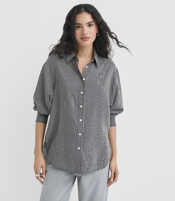 A woman with dark, wavy hair stands in the foreground wearing a black and white striped button-down shirt and light blue pants, against a plain white background.