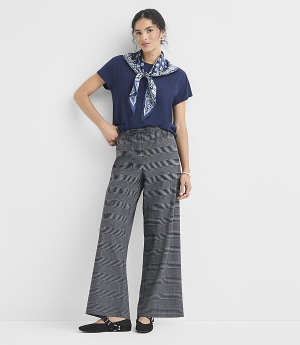 A woman in a navy t-shirt, patterned scarf, and plaid wide-leg pants stands against a plain white background.