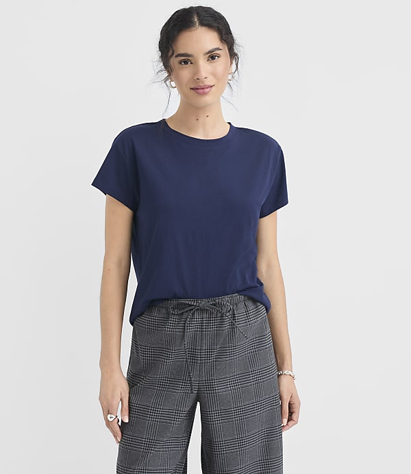 A woman in a navy t-shirt and plaid pants stands against a plain white background.