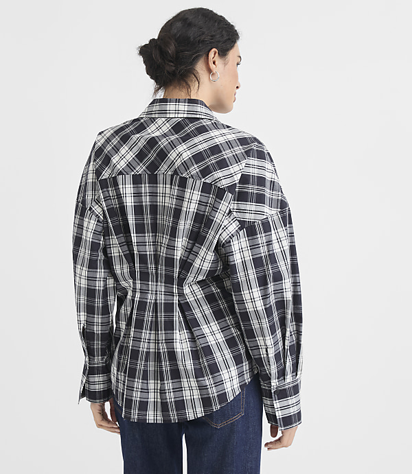A woman in a black and white plaid shirt stands against a plain white background.