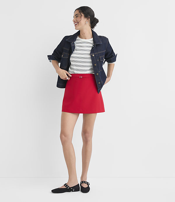 A woman in a denim jacket and red skirt stands against a plain white background.