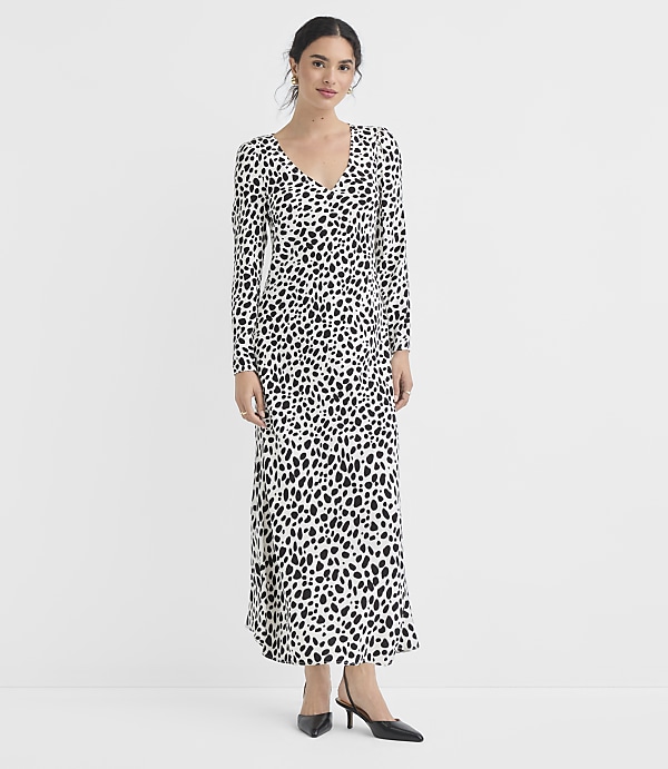 A woman in a long, black and white spotted dress stands against a plain white background.