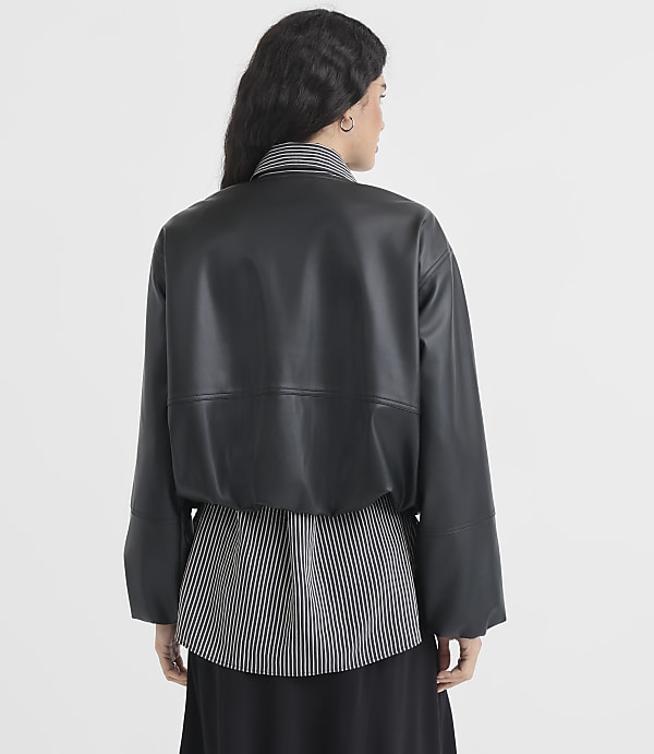A woman in a black faux leather jacket and striped shirt stands against a plain white background.