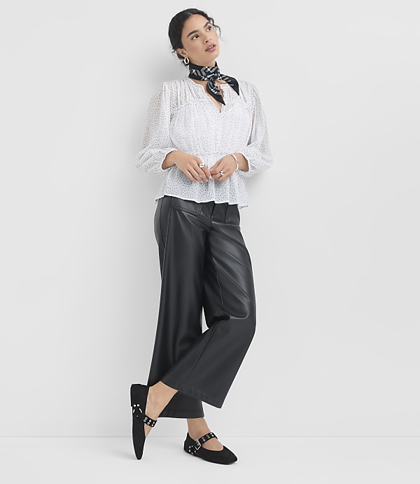A woman in a white polka-dot blouse and black leather pants stands against a plain white background.