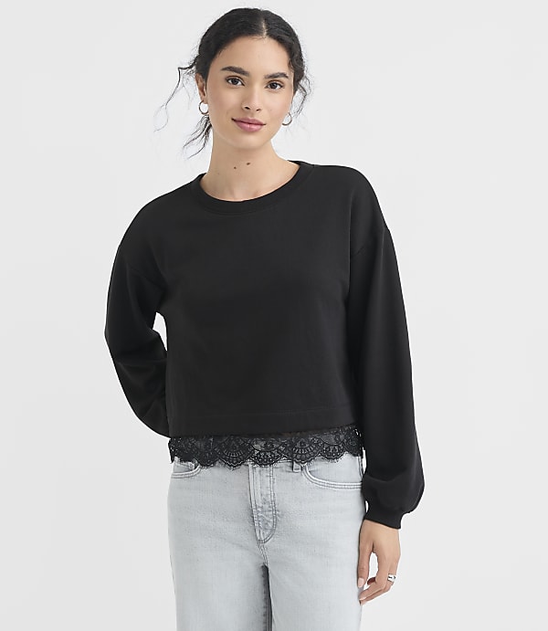A young woman in a black cropped sweatshirt with lace trim and light wash jeans stands against a plain white background.
