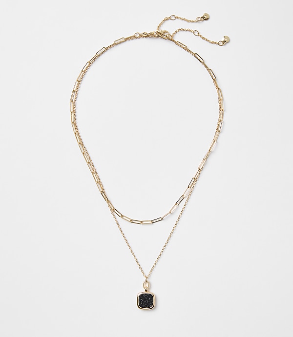A delicate gold layered necklace with a square black pendant rests on a plain white background.