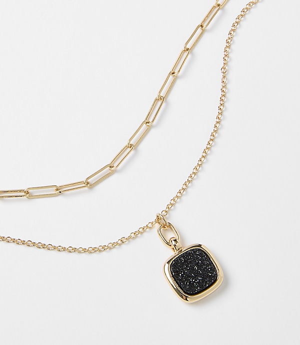 A layered gold necklace with a square black pendant hangs against a plain white background.