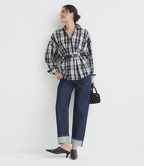 A woman in a plaid shirt and cuffed jeans stands against a plain white background, holding a small black handbag.