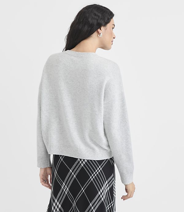 A woman in a light gray sweater and a black and white plaid skirt stands against a plain white background.