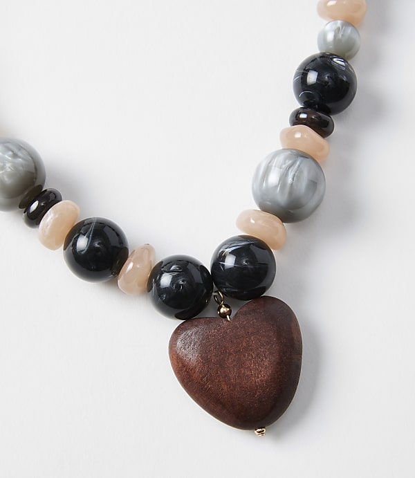 A wooden heart pendant hangs from a necklace of black, gray, and peach beads against a plain white background.