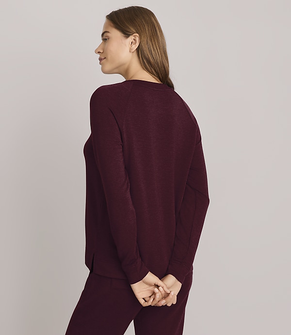 A woman in a purple sweatshirt and pants stands with her hands clasped behind her back against a plain gray background.