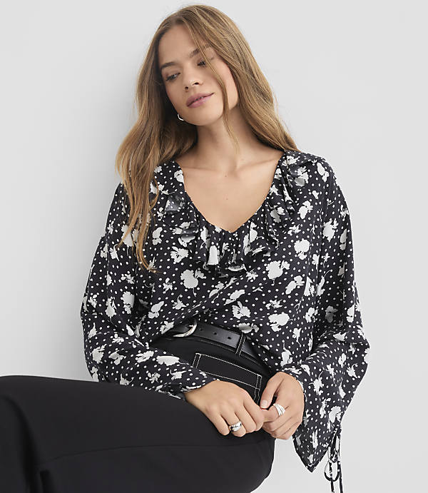 A woman in a black and white floral blouse with ruffled neckline and bell sleeves poses against a plain gray background.