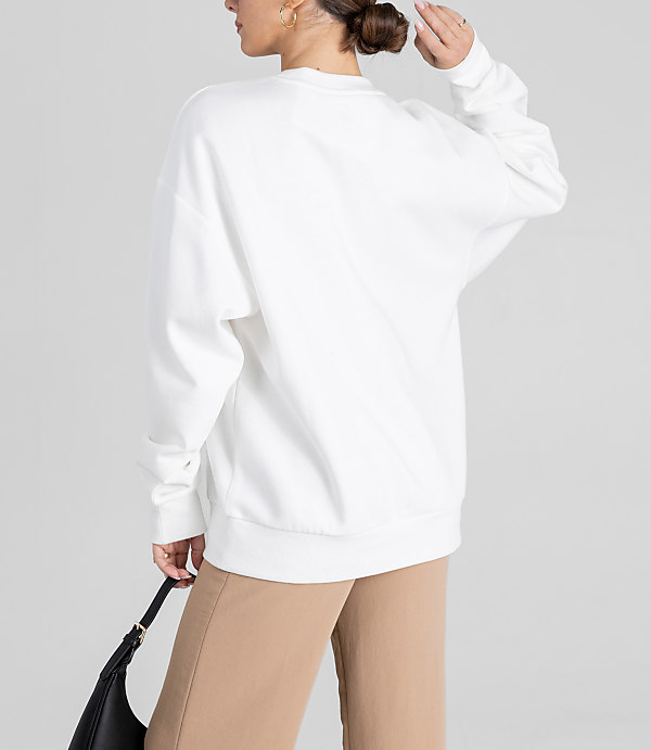 A woman in a white sweatshirt and tan pants holds a black purse against a plain gray background.
