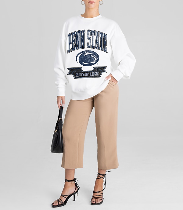 A woman in a white Penn State sweatshirt and tan cropped pants stands against a plain white background, holding a black purse and wearing black strappy heels.